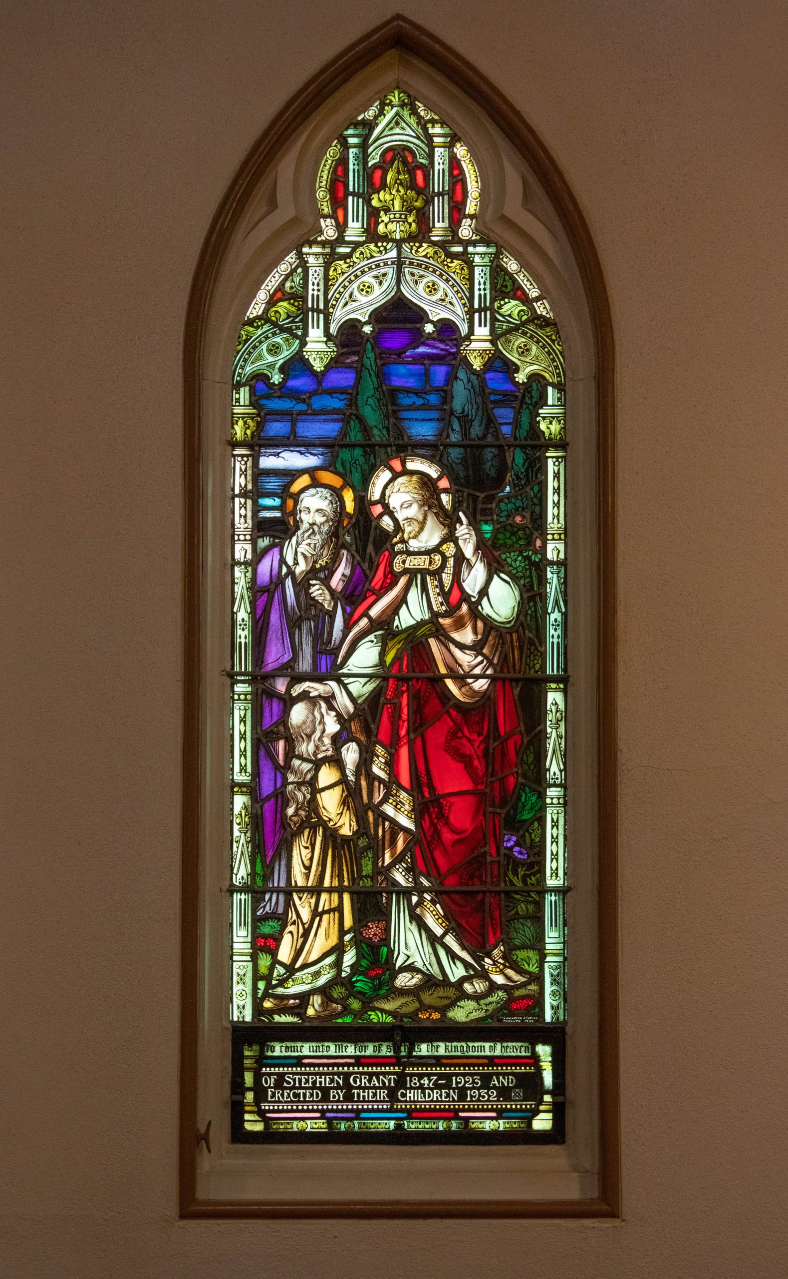 Stained glass window with two men and one child. One man lays hand on child's head in gesture of blessing.