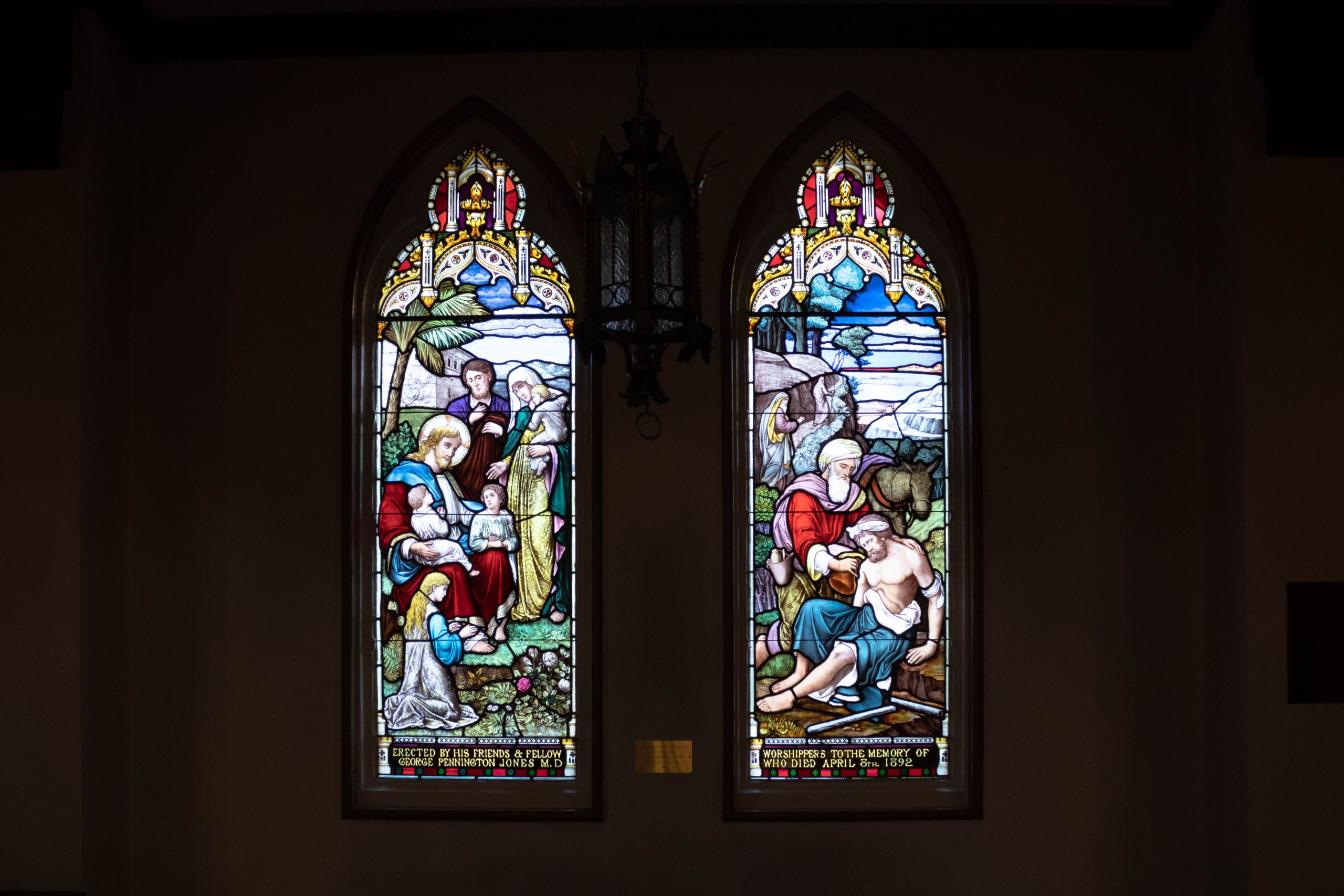 Two stained glass windows. Left window depicts adults and children. The right window shows a man helping another man.