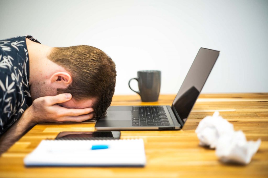 Image of man with head in hands, facedown on a desk. Laptop and office supplies around him.