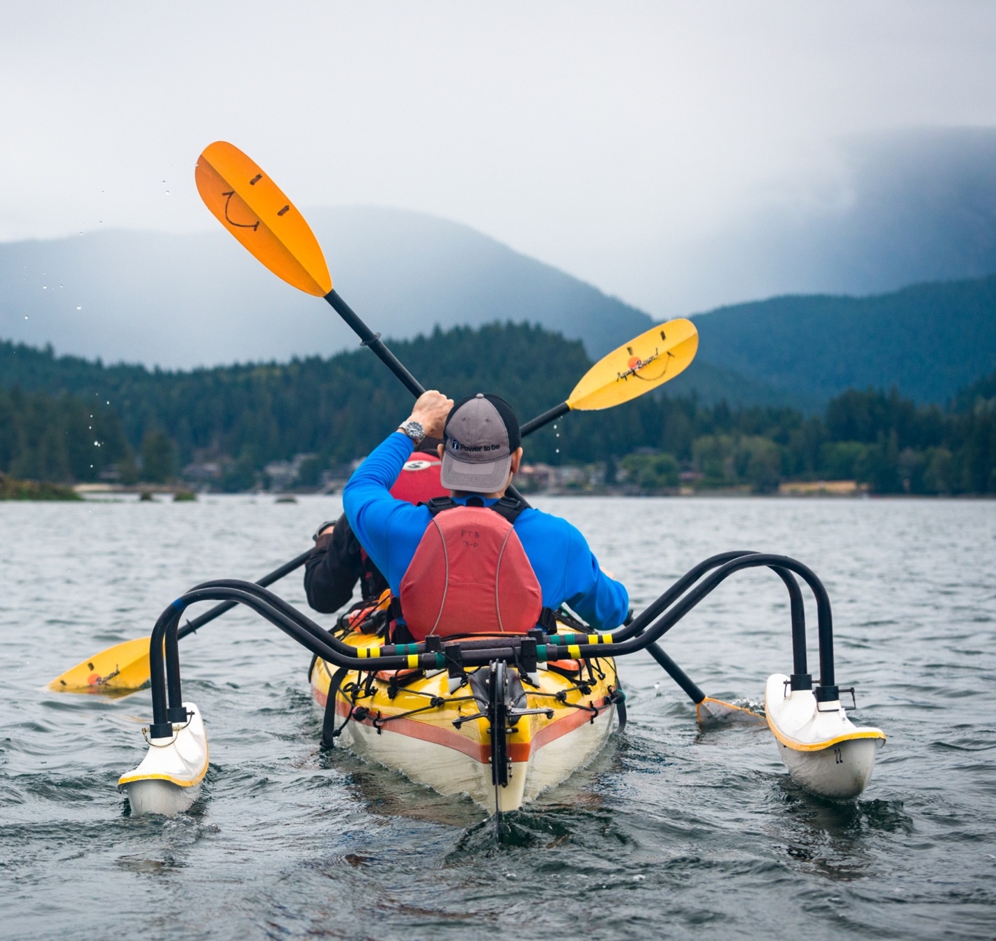 Two people in a kayak facing a mountain in the distance. The kayak has stabilizing pontoons.
