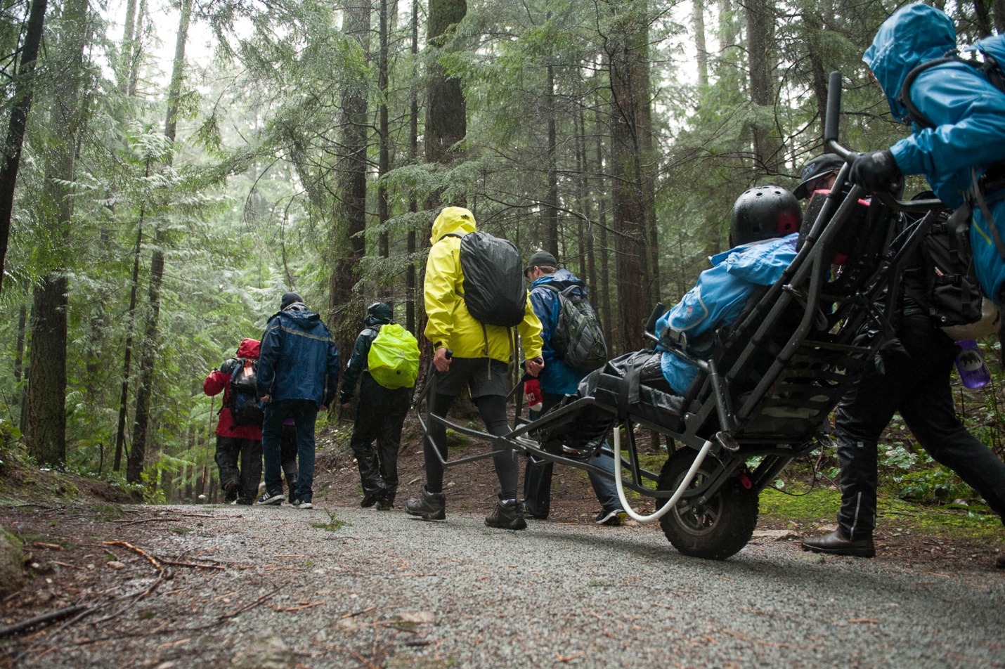 A group of people on a gravel hiking trail, with one person seated in a single-wheeled vehicle pulled by one hiker and steadied from behind by another