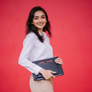 Young woman with white blouse, brown pants, medium length brown hair. She is holding a laptop.