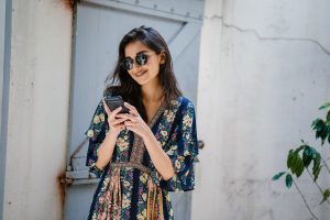 Young woman with shoulder length brown hair with a patterned dress and sunglasses. She is smiling and looking at her black phone.