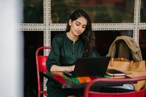 Young woman at a red table with a backpack on the chair beside her. She is wearing a green silk blouse and is typing on her laptop.