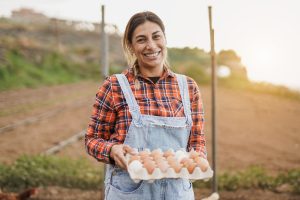 Middle aged woman in a field, wearing overalls and a plaid shirt, holding a tray of white and brown eggs.