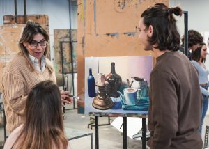 Three people in an art studio with one middle-aged women with glasses speaking to two other people who are facing a painting.