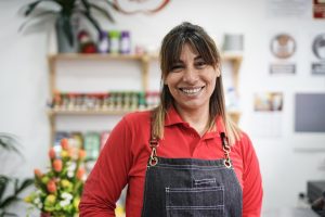 Photo of a middle-aged woman smiling in a food store, wearing overalls and a red shirt.