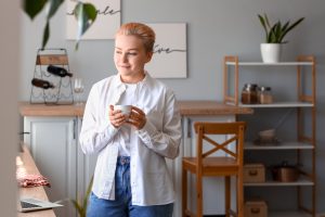 Young woman with short blonde hair standing in an apartment. The women has a white button up shirt and jeans and is holding a white coffee cup.