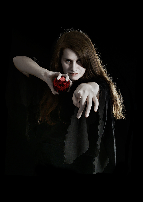 A female with pale skin dressed in black holding a red ball and pointing down the camera lens