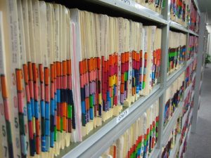 Close-up on rows of organized file folders on metal shelves within what appears to be a records storage area. The folders are vertical, with tabs in red, blue, orange, yellow, and green and primary digits moving from 30 at left to 31 in the centre, and 34 on the lower shelf.