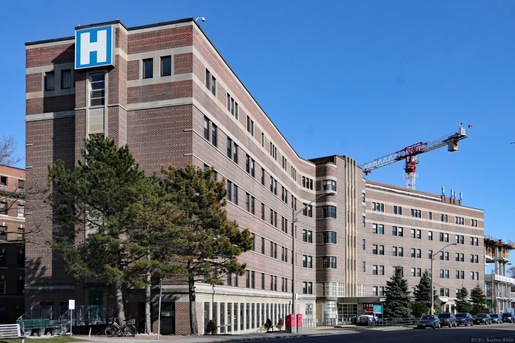 A mid-20th-century red brick hospital building with a blue "H" sign and a tree in the foreground extends horizontally across the image with a slight curve, suggesting two wings on either side of the centre. The sky is bright blue and there is a construction crane in the right background.