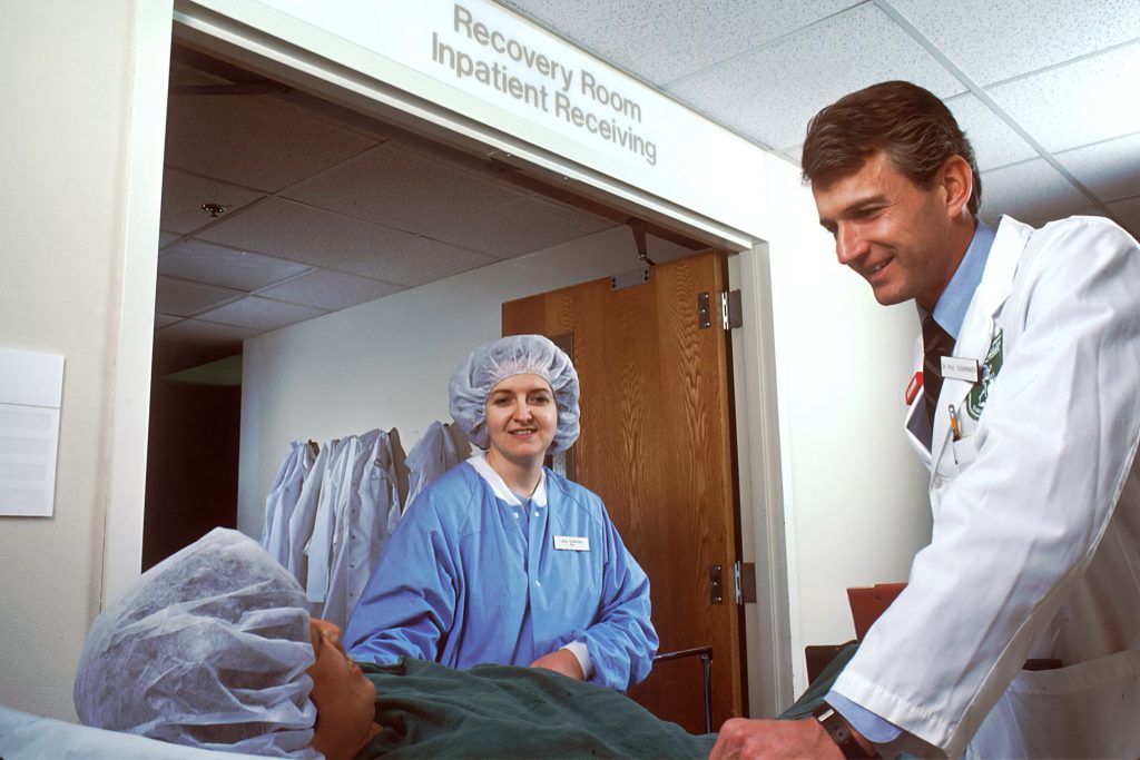 A photo of a patient on a gurney, taken at lower than eye level, just outside the surgery recovery room; on either side of gurney a smiling woman in blue surgical scrubs and smiling man in doctor's lab coat and shirt and tie look down toward at patient.