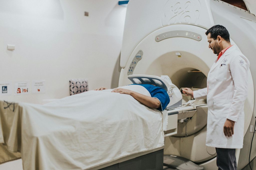 A patient lying on a platform covered with a white sheet, about to enter the large cylindrical opening of an MRI machine head-first. To the right of the patient, a male healthcare professional, dressed in a white lab coat is interacting with the patient.