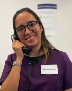 A young woman in purple scrubs and a nametag that reads "Health Care Administrator" is holding the receiver of a telephone and smiling at the camera.