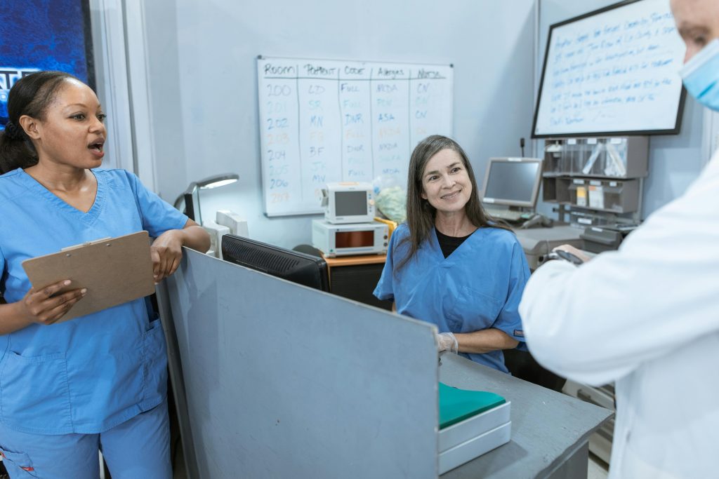 Two women in scrubs and a person in white coat have a discussion around a small nursing station with a whiteboard on the wall that lists room number, patient initials, code (such as DNR), allergies, and notes.