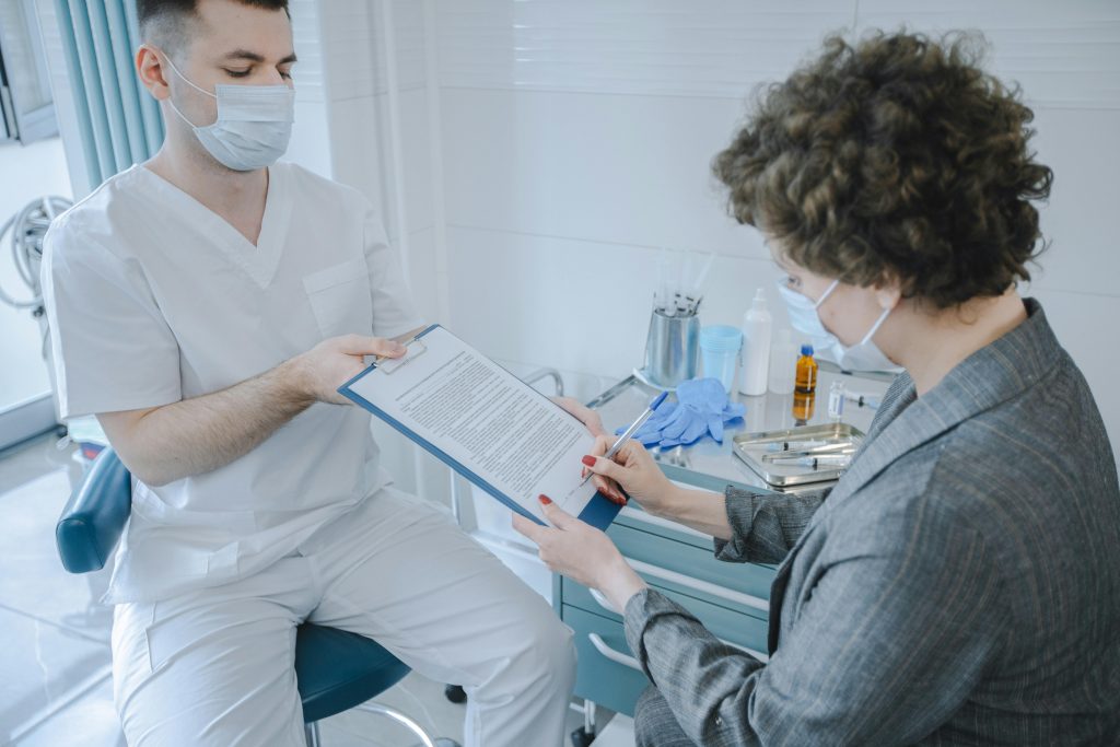 A healthcare professional in a clinical setting wearing white scrubs and a face mask sitting on a stool, holding a clipboard for a patient wearing a gray blazer who is signing the document.