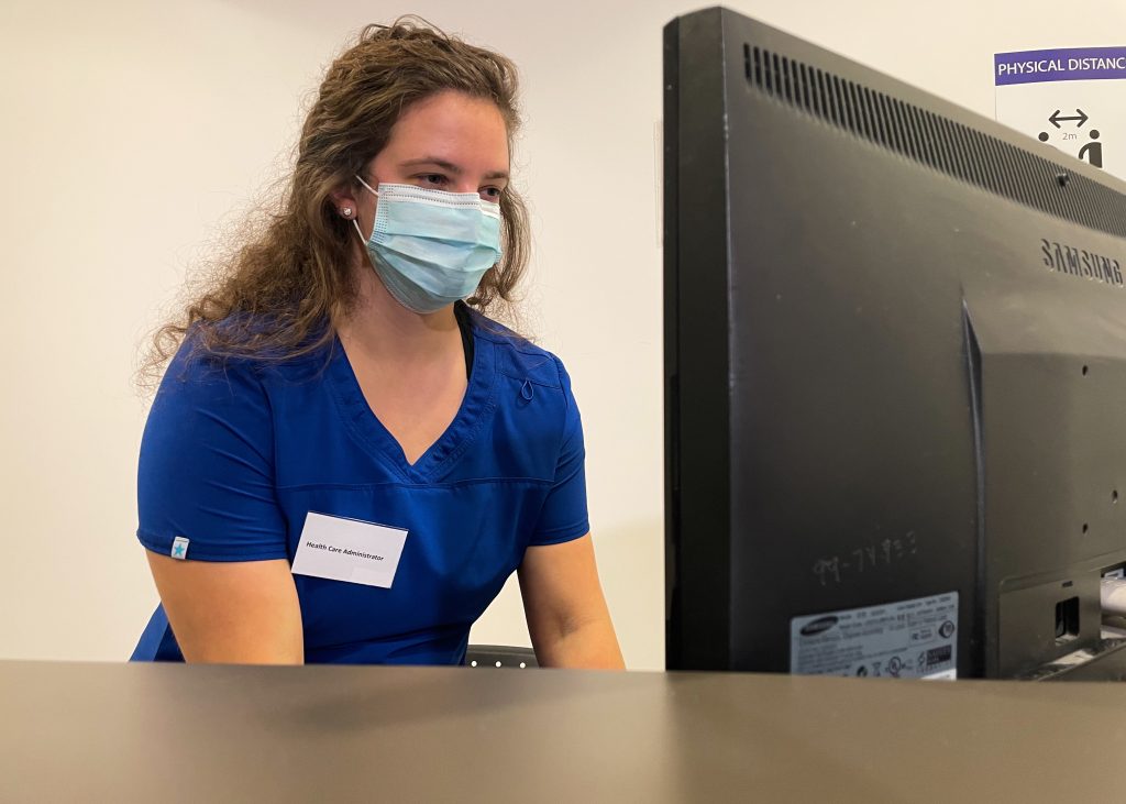 A woman in blue scrubs, facemask, and a nametag that reads "Health Care Administrator" behind a high desk is looking at a computer display and leaning forward with arms slightly in front, suggesting she is typing below the level of the desk.