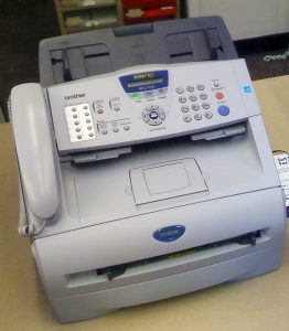 Close-up of a Brother fax machine viewed from above on a desk, which looks like a small desktop printer with a panel of operational buttons on the top; there is a phone receiver on the left of the unit and a number key pad on the right side of the control panel.