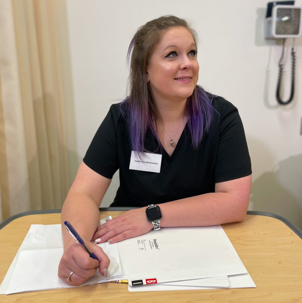 A woman in dark blue scrubs with a nametag that reads "Health Care Administrator" is seated at a table in an exam area, writing on paper in a file folder while looking up towards someone off camera.