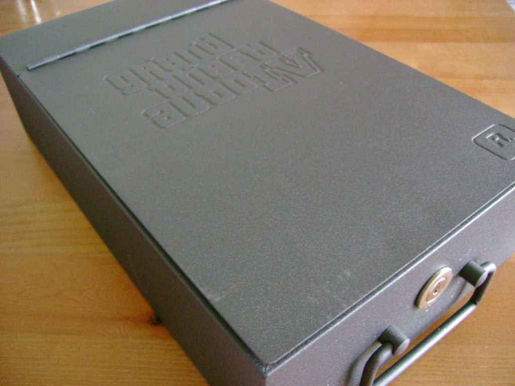 Close-up of a sturdy gray metallic rectangular safety deposit box that is several inches long, half as wide but only a few inches high; the box opens at a hinge on the lid that is a few inches from the far edge. A metal handle is on the front side, with a round gold lock on the box.