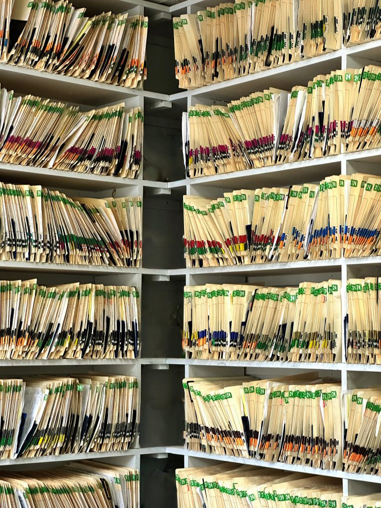 Shelves filled with neatly aligned manila file folders, labelled at the top edge with tabs of various colours, predominantly green, with some showing numbers and letters; the orderly arrangement suggests an efficient filing system, possibly in a records room.