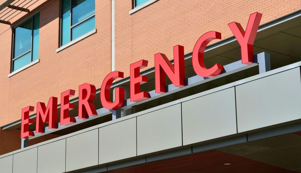 A close-up of a hospital building exterior with the word "EMERGENCY" across the middle of the image in individual red letters attached to a bar above what appears to be the covered entrance area, which is not pictured.