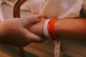 Close-up of a person's arm resting on a hospital bed rail being held by a another person’s hand. The arm has two identification wristbands, one orange and one white. The orange wristband has the word "ALLERGY" visible.