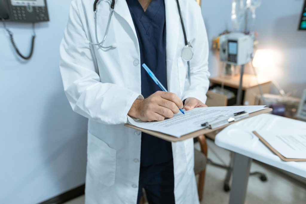 Close-up of a physician (torso and arms only) in scrubs, white coat, and stethoscope making notes on a clipboard in a patient room.