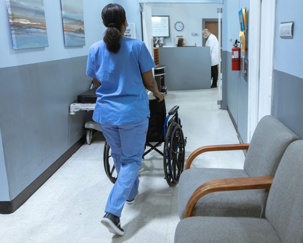 A woman in blue scrubs walking away from the camera pushes an empty wheelchair down a narrow hospital hallway towards a nursing station.