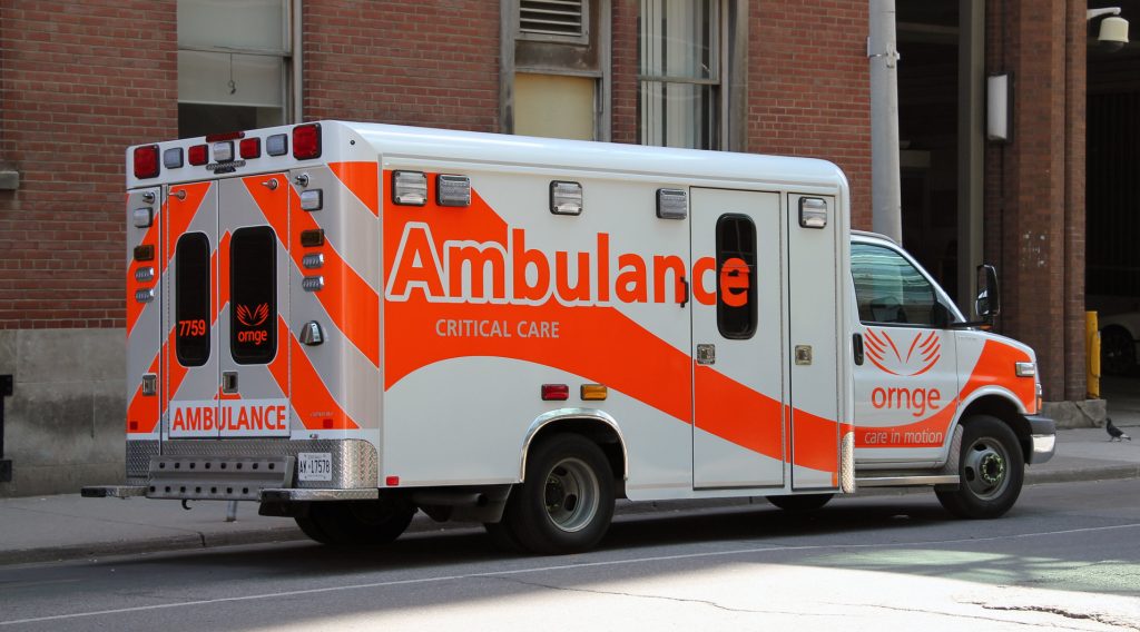 A white and orange ambulance parked on a side street next to a red brick wall with windows. "Ambulance" is written on the side of the vehicle, and the ORNGE logo and wordmark are on the passenger side cab door.