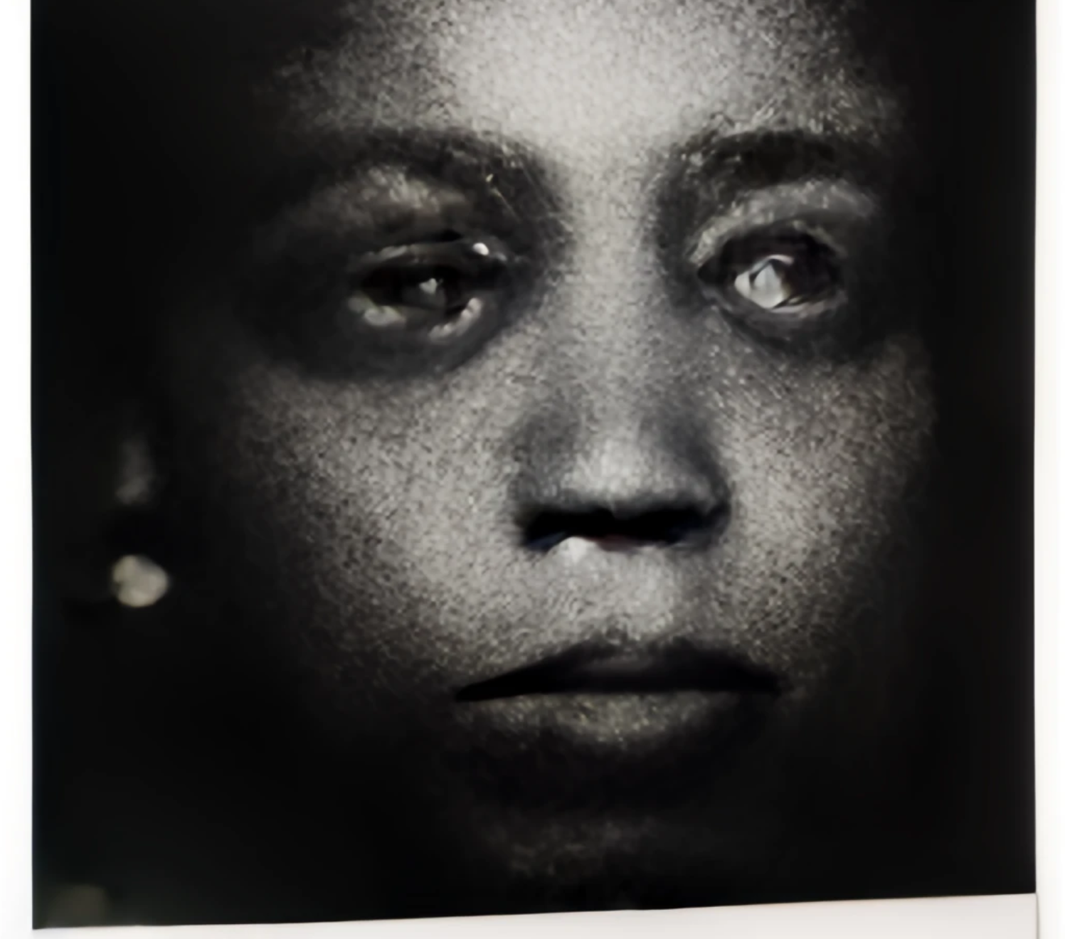 Black and white photo, with a thin slanted white border around the sides and bottom. Pictured is a youthful face, lit from above but receding into the darkness. They have full eyebrows, dark eyes, a flat, wide nose, and full, downturned lips. Their expression is stoic, but their eyes are weltered by the AI generator, giving them a tearful, swollen appearance. Although their eyes are marred, their gaze lands distinctly to the views right. Their skin has a cross hatched or scales texture. An earring in their right ear is dimly visible.