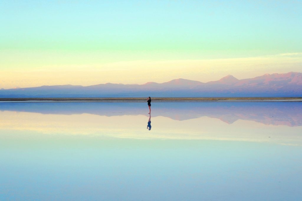 Silhouette of a person standing in a shallow reflective lake with distant mountains and a blue sky at sunset.