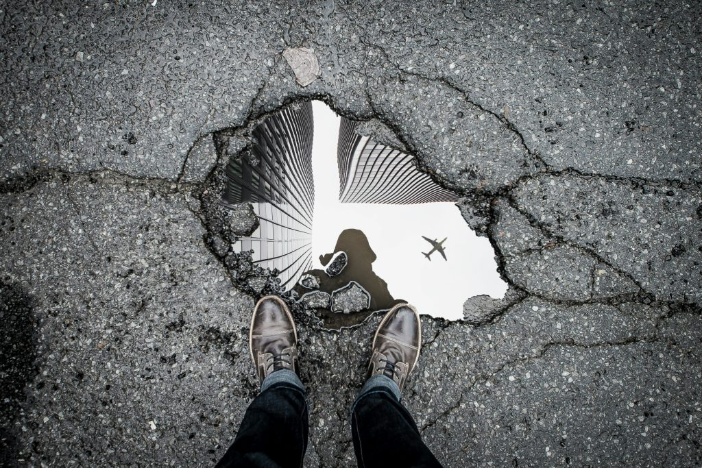 A person standing beside a pothole filled with water reflecting a building and an airplane flying overhead, as seen from their point of view.