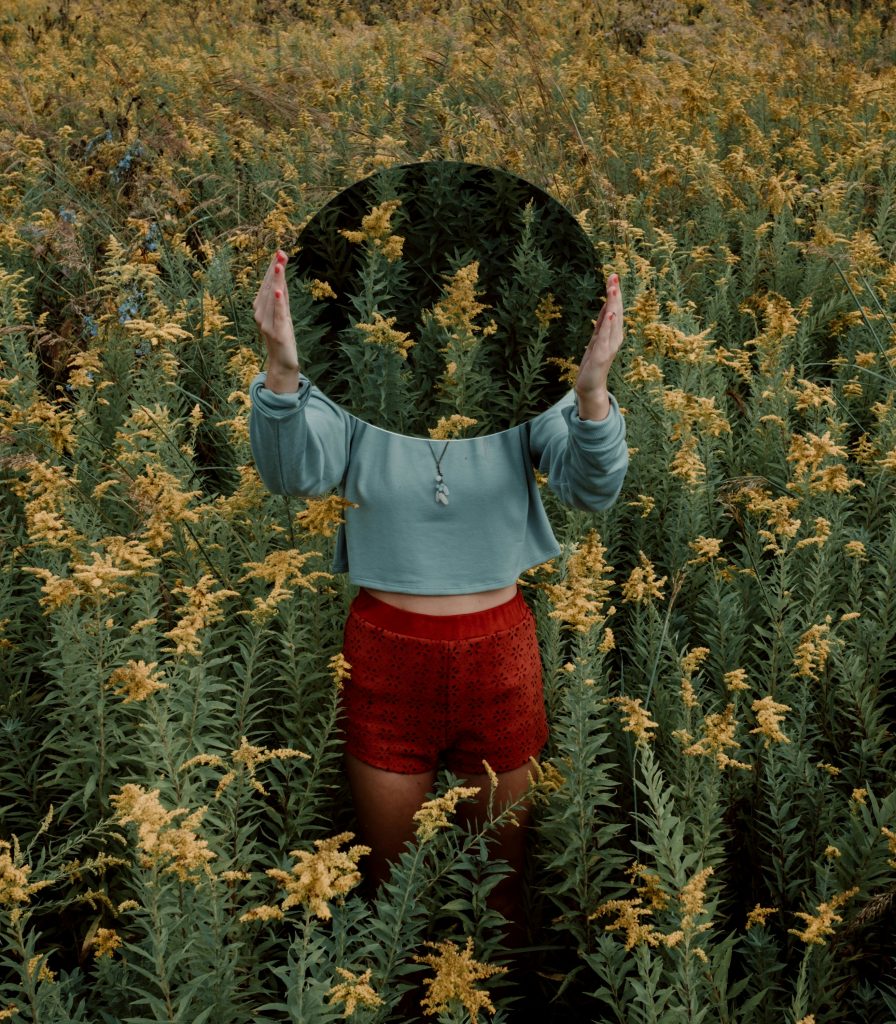 A person holds a circular mirror in front of their face, surrounded by tall yellow wildflowers.