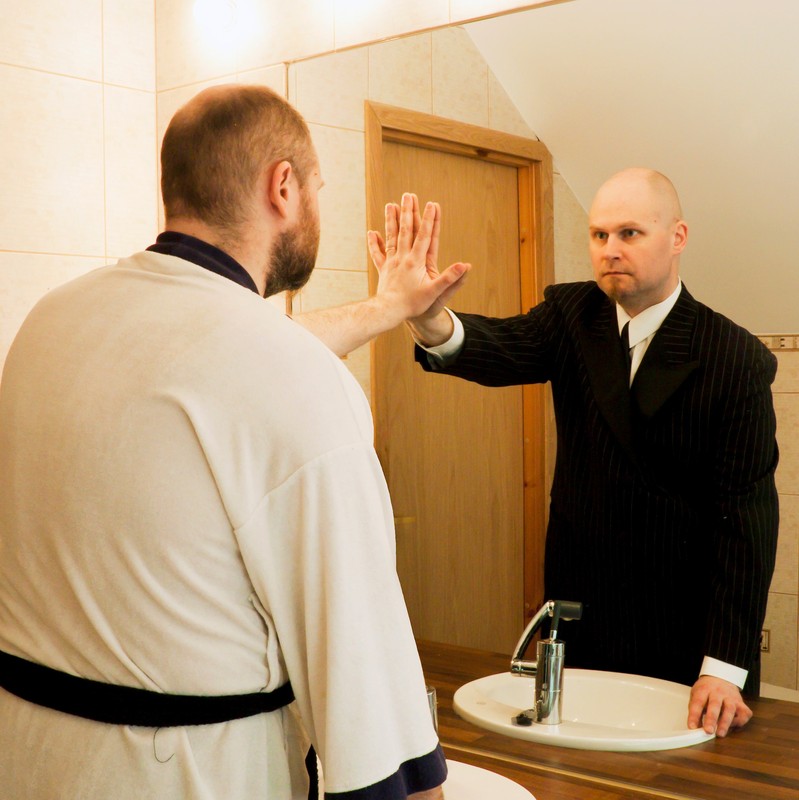 Photo of a man looking at himself in a mirror. He is wearing a bathrobe and his mirror image is wearing a suit.