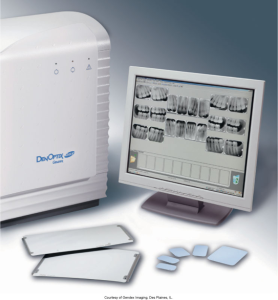 A DenOptix QST Storage Phosphor Imaging System next to a computer monitor displaying dental X-ray images, with assorted imaging plates on the table, courtesy of Gendex Imaging, Des Plaines, IL.