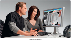 A dentist and a patient looking at a dental X-ray displayed on a computer monitor, with specific areas of concern highlighted in red circles, courtesy of DEXIS, Des Plaines, IL.