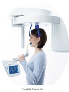 An image of a woman undergoing a dental X-ray examination. She is positioned within a panoramic dental X-ray machine, using a head stabilizer and bite block for accurate imaging. The control panel of the machine is visible in the foreground, indicating the interactive components for operating the X-ray system.