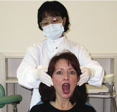 A dentist wearing a mask and gloves is examining a female patient's open mouth, checking for temporomandibular disorder in a clinical setting.