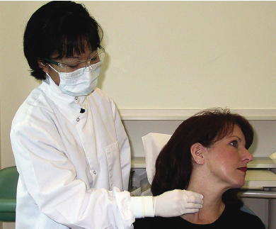 A healthcare professional wearing a lab coat, mask, and eye protection is examining a female patient's neck for palpation of the sternocleidomastoid muscle.