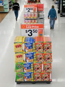 Four columns and four rows of cereal boxes stacked at the end of a supermarket's aisle with a large "low price" sign across the top.