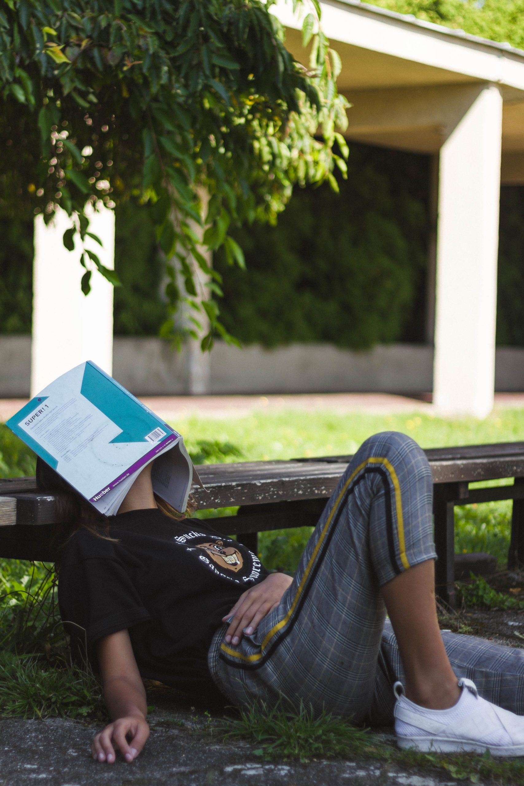 Decorative photograph of a tired student laying down in the park with an opened book covering their face.