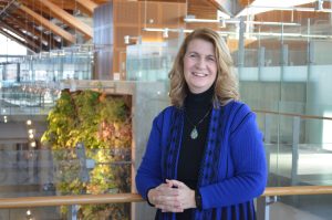 Lorie Stolarchuk in the Centre for Engineering Innovation at the University of Windsor. Wearing a black turtleneck and black and blue sweater. Plants in the background
