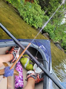 a blue kayak sits in quiet water near a shore. The person in the kayak is spinning on a cross-bar spindle, while their paddle lies across the kayak