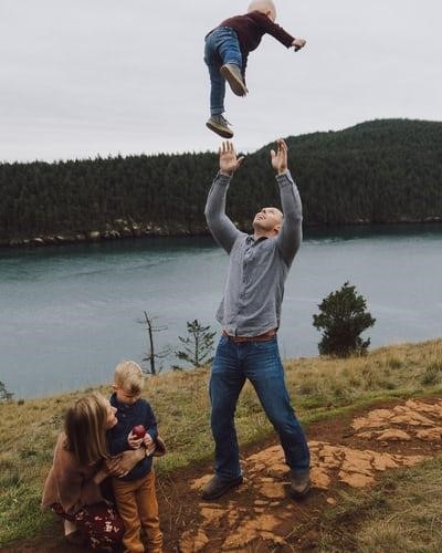 mother, father and 2 young children standing by a body of water. The father is tossing one of the children up in the air. The mother is kneeling down with her arms around the other child.