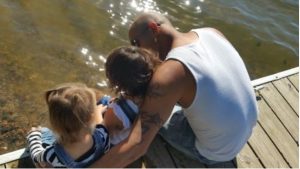 A father sitting on a dock with his arms around his two young children.