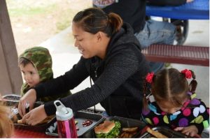 A mother sitting at a pcnic table with her two young children, helping them eat their lunch.