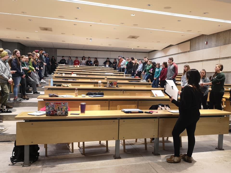 Photograph of students standing in a lecture hall all facing each other in a circle