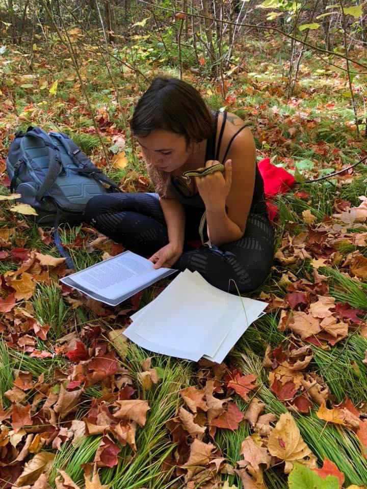 female student sitting on the forest floor with a snake in her hand while reading a stack of papers