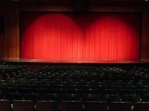 An image of an empty stage with a spotlight shining on red theater curtains.